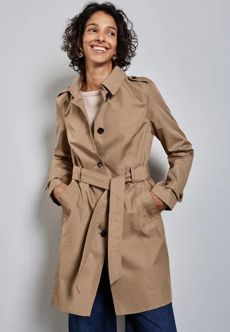 Lächelnde Frau im beigefarbenen Trenchcoat mit Gürtel vor grauem Hintergrund.